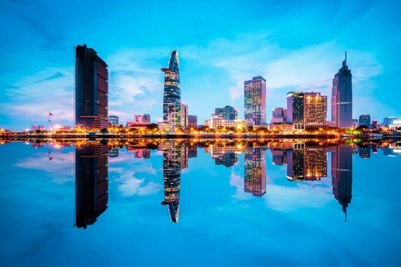 Cityscape In Reflection Of Ho Chi Minh City At Beautiful Twilight, Viewed Over Saigon River. Hochiminh City Is The Largest City In Vietnam With Population Around 10 Million People